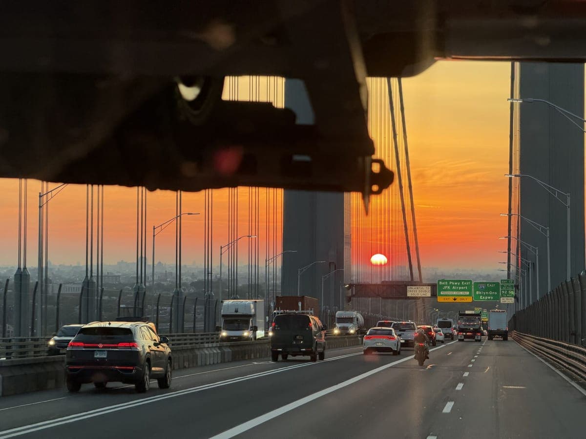 Sunrise seen from a truck cab crossing the Verrazano-Narrows Bridge toward Brooklyn, with traffic and highway signs ahead.