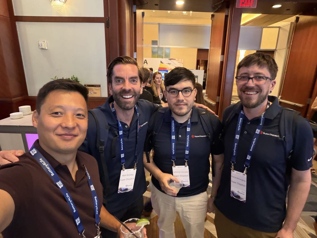 Four attendees wearing conference lanyards smiling together in a selfie at an industry event.